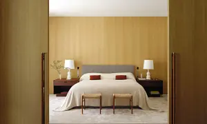 Camera da letto con letto centrale, testiera grigia, comodini in legno scuro e due sgabelli. Ottima idea su come posizionare il letto in camera per un look armonioso.