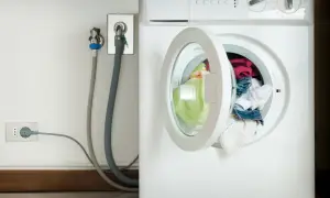 Lavatrice con oblò aperto e panni colorati. Vicino, il rubinetto per l'acqua e la presa elettrica, con un tubo grigio che collega il tutto. L'altezza del rubinetto lavatrice è standard.