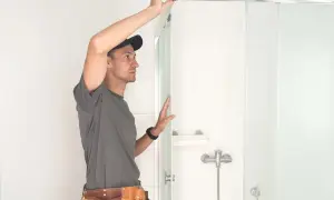 Operaio con cintura porta attrezzi impegnato nel montaggio box doccia, con cura posiziona un pannello di vetro.