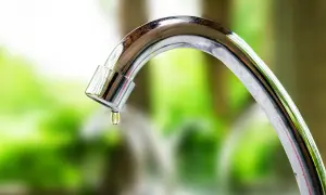 Goccia d'acqua cade da un rubinetto che perde. La manopola del rubinetto è lucida e riflette il verde dello sfondo.