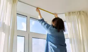 Persona misura l'altezza del bastone per tende dal soffitto, con tende bianche e una finestra sullo sfondo.