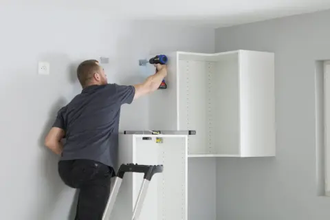 Uomo su scala avvita staffe per come appendere i pensili della cucina. Livella a bolla posizionata.