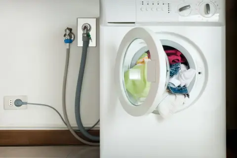 Lavatrice con oblò aperto e panni colorati. Vicino, il rubinetto per l'acqua e la presa elettrica, con un tubo grigio che collega il tutto. L'altezza del rubinetto lavatrice è standard.