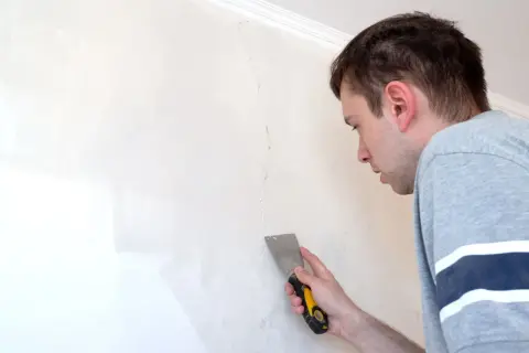 Uomo con spatola ripara una crepa su un muro scrostato, pronto a stuccare.