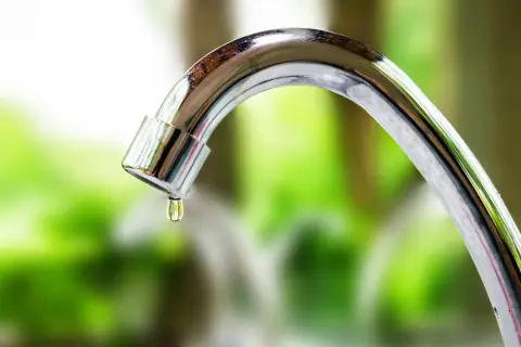 Goccia d'acqua cade da un rubinetto che perde. La manopola del rubinetto è lucida e riflette il verde dello sfondo.