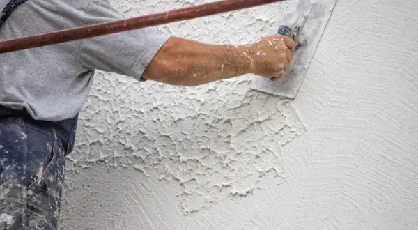 Mano di un operaio che applica la finitura intonaco rustico con una spatola, creando una texture irregolare e caratteristica.