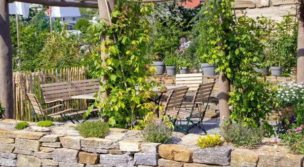 Un rigoglioso giardino con un muro a secco per contenimento terra, tavoli e sedie in legno sotto un pergolato.