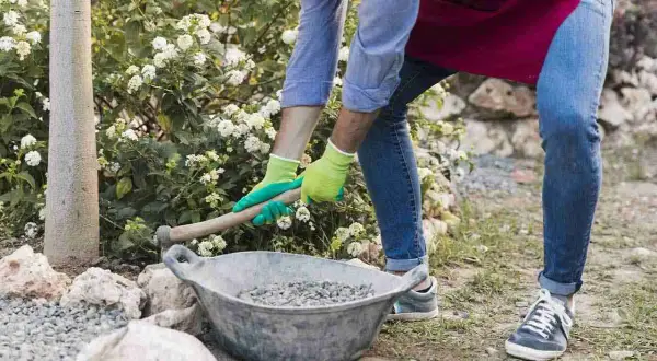 Uomo con guanti verdi prepara la ghiaia per una gettata di cemento in giardino, vicino a un albero e fiori bianchi.