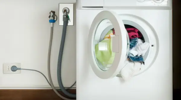 Lavatrice con oblò aperto e panni colorati. Vicino, il rubinetto per l'acqua e la presa elettrica, con un tubo grigio che collega il tutto. L'altezza del rubinetto lavatrice è standard.