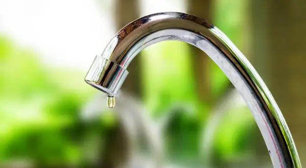 Goccia d'acqua cade da un rubinetto che perde. La manopola del rubinetto è lucida e riflette il verde dello sfondo.
