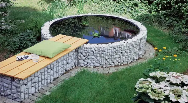 Un incantevole laghetto fuori terra fai da te, circondato da pietre bianche e con una panchina in legno per rilassarsi.