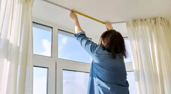 Persona misura l'altezza del bastone per tende dal soffitto, con tende bianche e una finestra sullo sfondo.