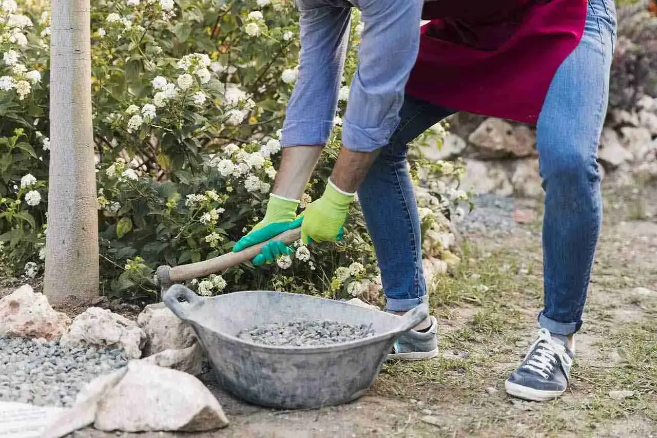 Uomo con guanti verdi prepara la ghiaia per una gettata di cemento in giardino, vicino a un albero e fiori bianchi.