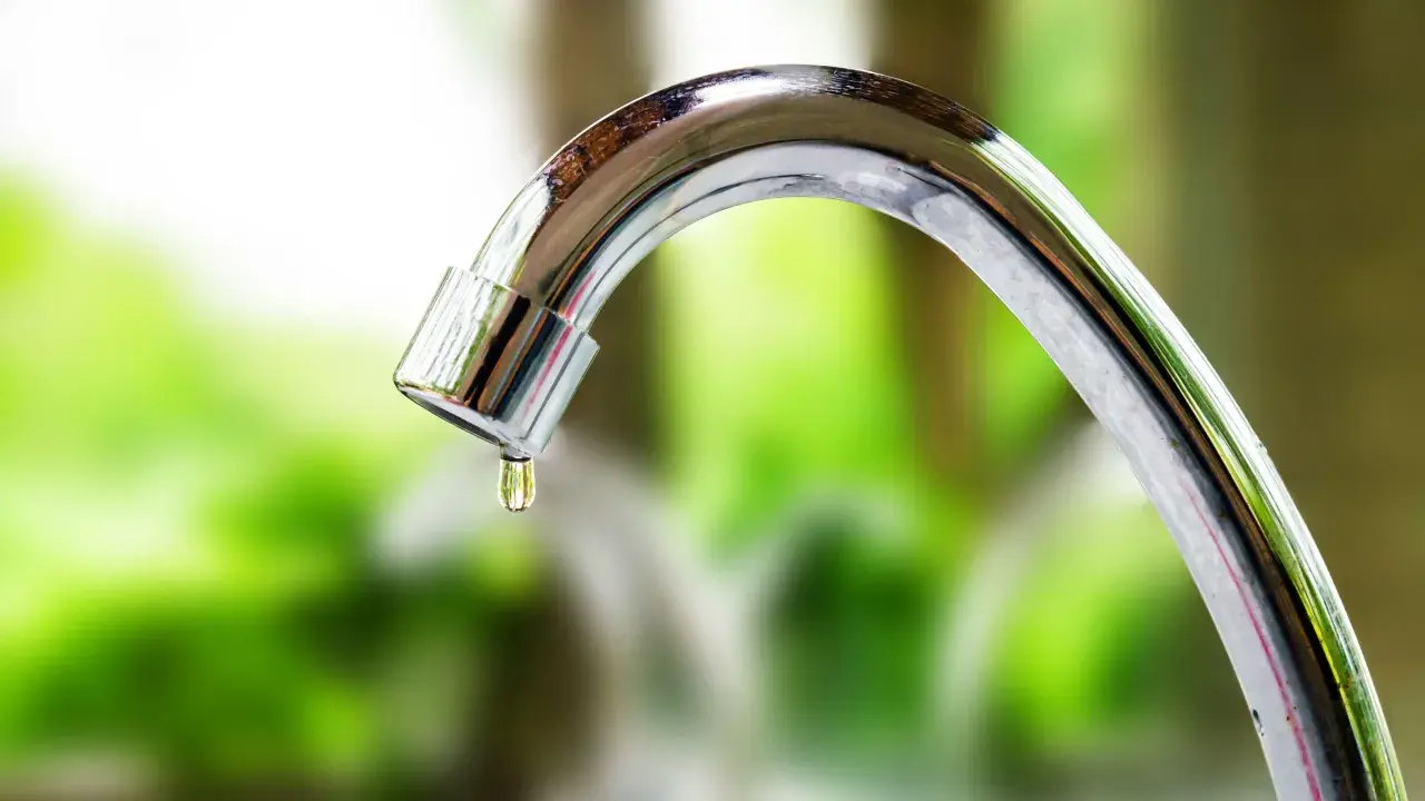Goccia d'acqua cade da un rubinetto che perde. La manopola del rubinetto è lucida e riflette il verde dello sfondo.