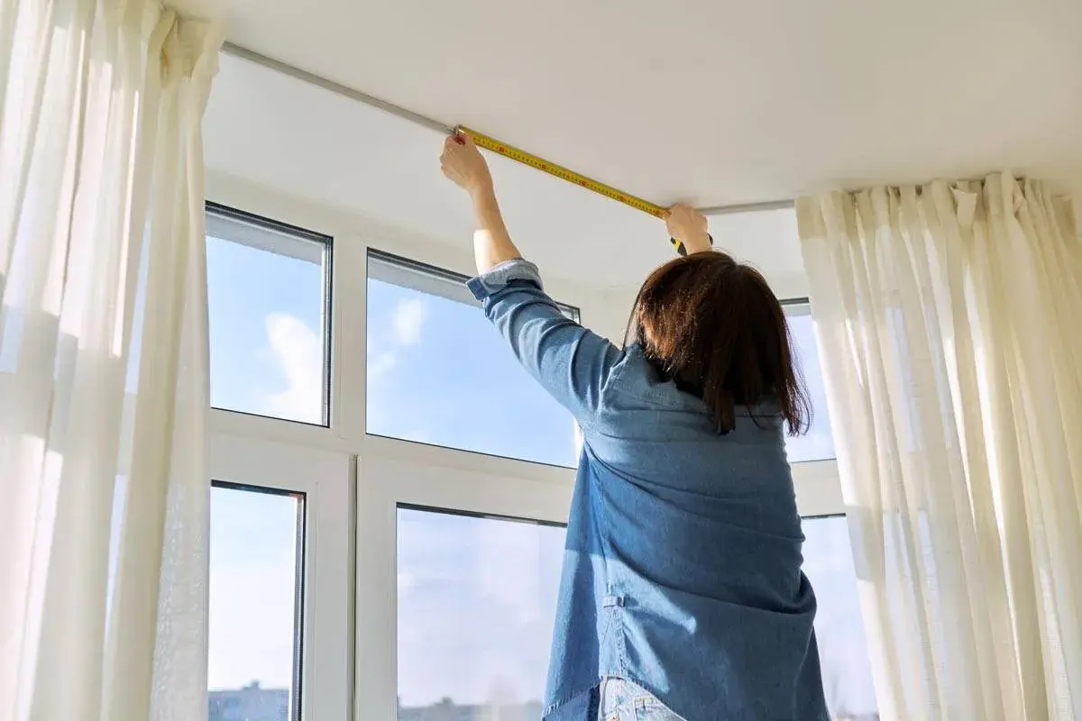 Persona misura l'altezza del bastone per tende dal soffitto, con tende bianche e una finestra sullo sfondo.