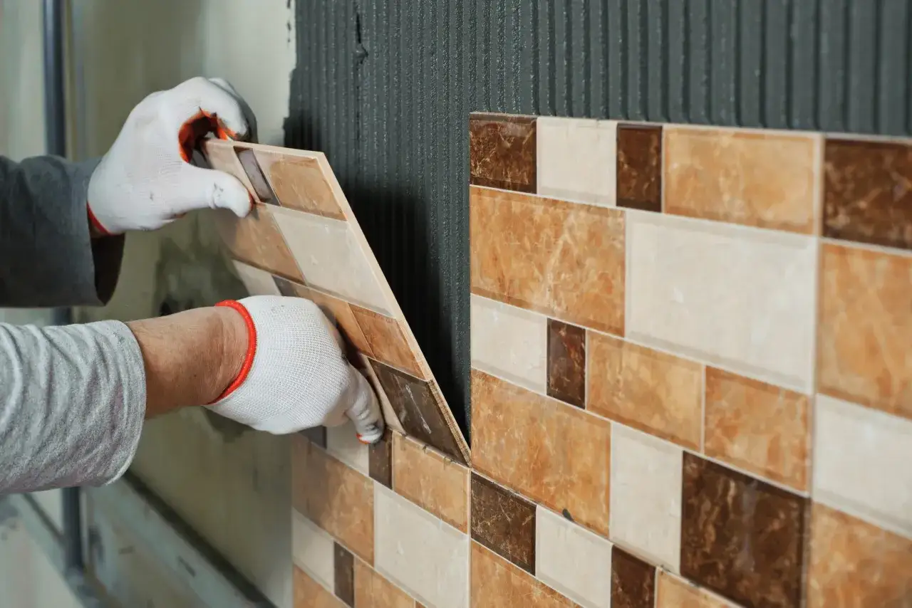 Mani guantate applicano una piastrella beige e marrone su muro con colla, mostrando come posare le piastrelle del bagno.