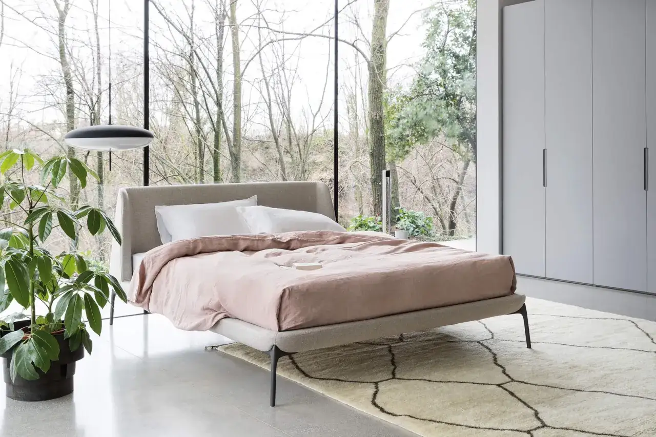 Un letto moderno con lenzuola rosa cipria e cuscini bianchi, un vaso con pianta verde e un lampadario minimalista. Ideale per chi cerca cosa mettere davanti al letto per un tocco di stile.