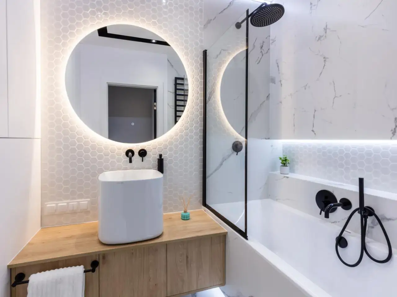 Moderno bagno cieco idee: lavabo bianco su mobile in legno, specchio rotondo illuminato, vasca da bagno con doccetta nera.
