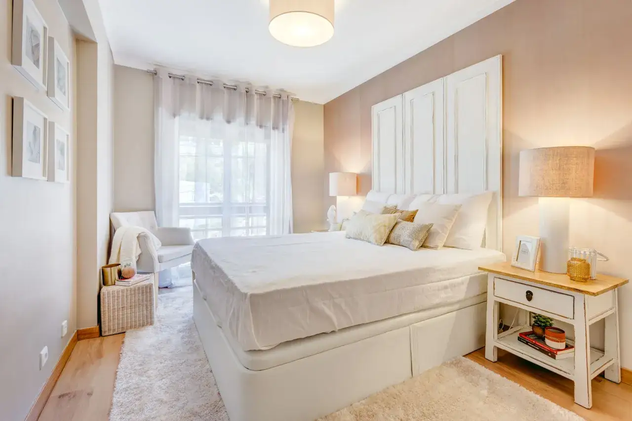 Ecco un esempio di come posizionare il letto in camera: un letto bianco con testiera decorata, comodini, poltrona e tappeto soffice creano un'atmosfera rilassante.