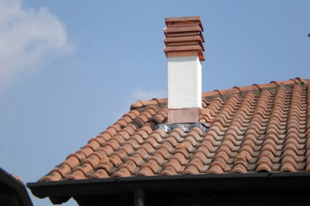 Manutenzione canna fumaria privata in condominio: un comignolo bianco con cappello in rame su tetto di tegole rosse, sotto un cielo azzurro con nuvole.
