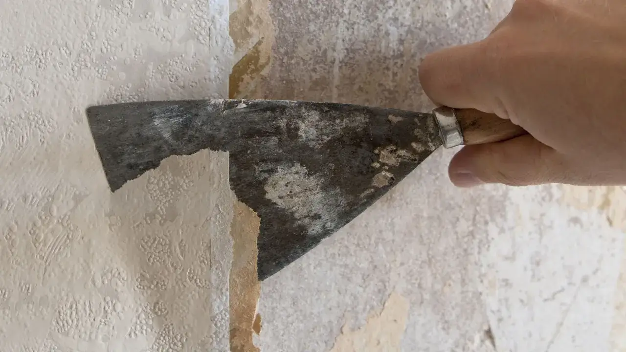 Mano con spatola rimuove vecchia carta da parati, un passo fondamentale per sapere come mettere carta da parati nuova.