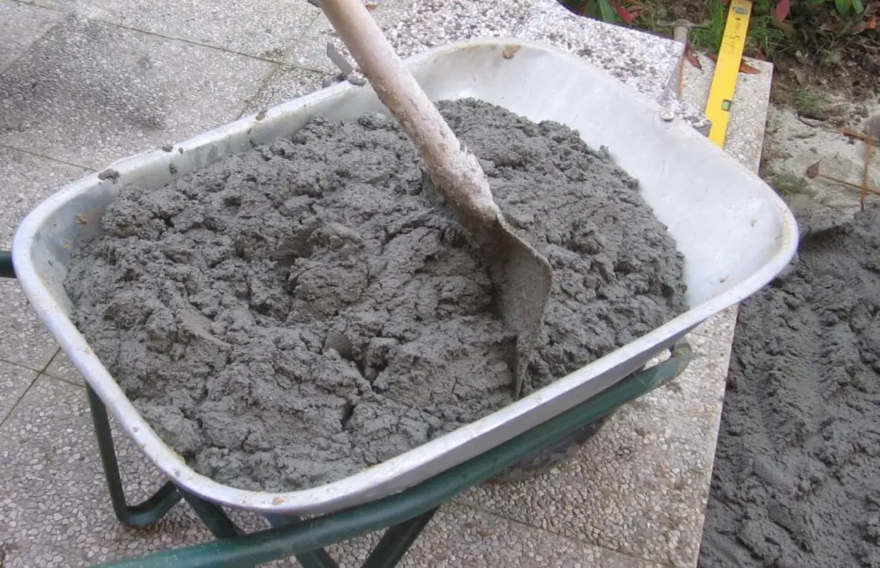 Carriola piena di cemento fresco, pronto per essere utilizzato. Si vede una pala che mescola il composto, mostrando come si fa il cemento per lavori edili.