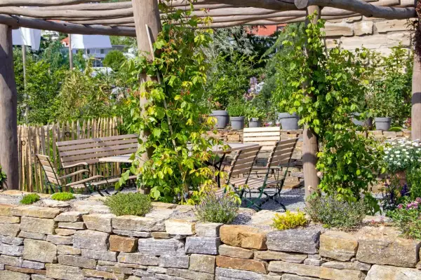 Un rigoglioso giardino con un muro a secco per contenimento terra, tavoli e sedie in legno sotto un pergolato.