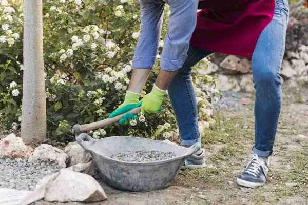 Uomo con guanti verdi prepara la ghiaia per una gettata di cemento in giardino, vicino a un albero e fiori bianchi.
