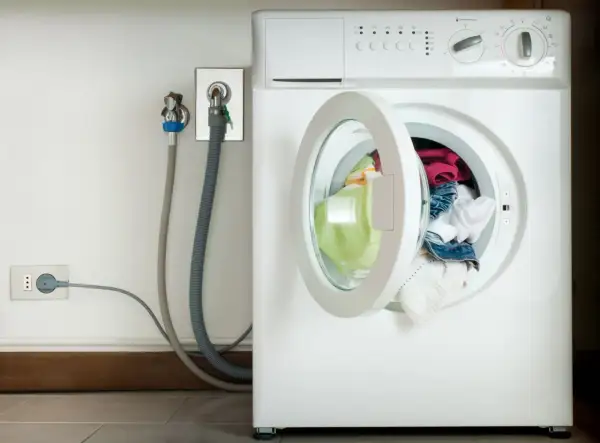 Lavatrice con oblò aperto e panni colorati. Vicino, il rubinetto per l'acqua e la presa elettrica, con un tubo grigio che collega il tutto. L'altezza del rubinetto lavatrice è standard.