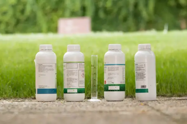 Bottiglie di prodotti agricoli, tra cui un erbicida selettivo, e un cilindro graduato su un vialetto di ghiaia, con prato verde sullo sfondo.