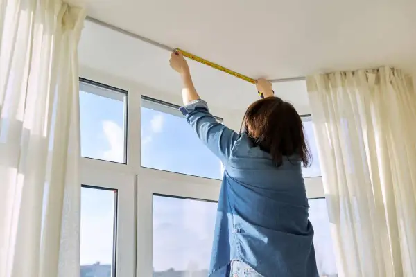 Persona misura l'altezza del bastone per tende dal soffitto, con tende bianche e una finestra sullo sfondo.